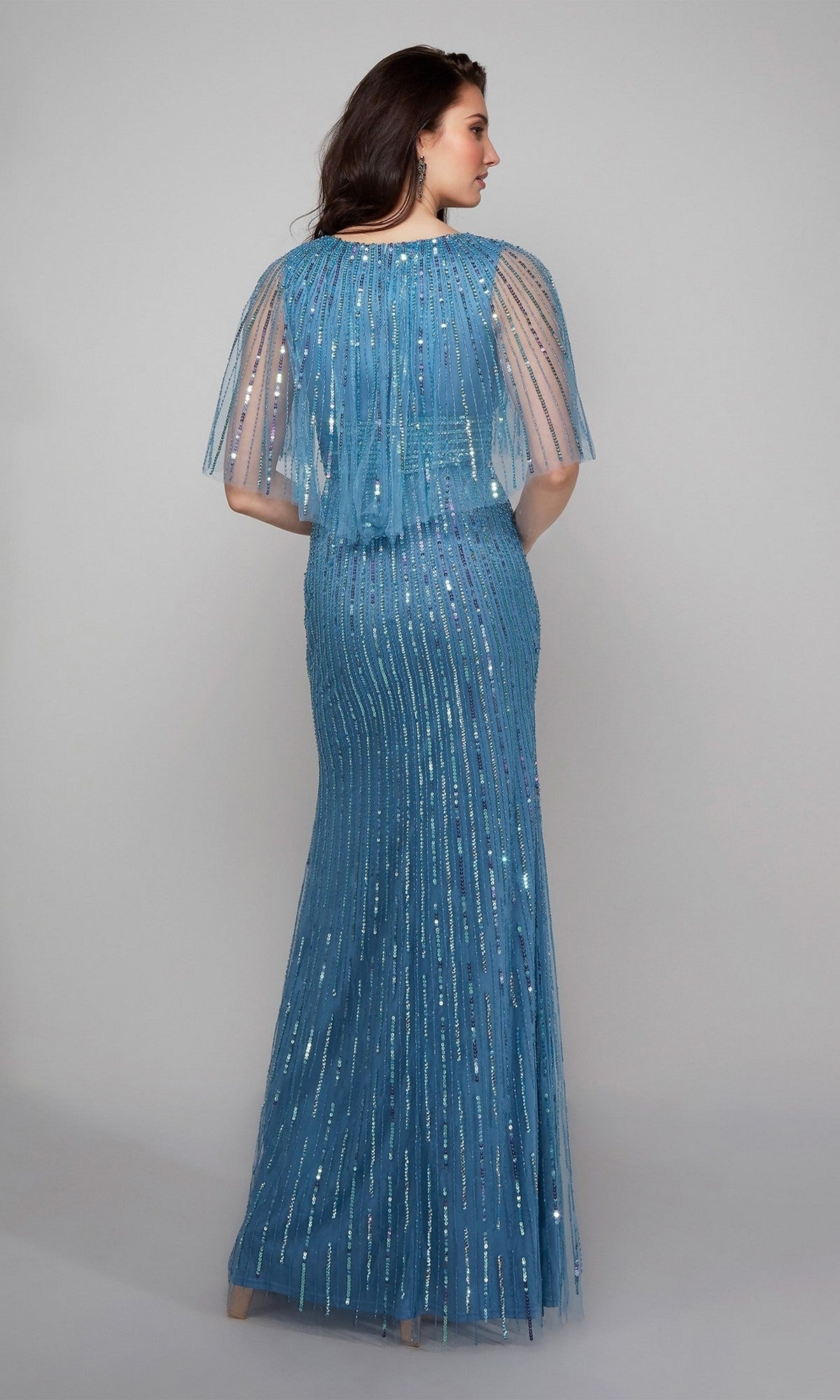 Woman wearing the Alyce 27599 long formal dress—floor-length, fitted, blue with sheer beaded overlay and short cape sleeves—stands against a plain background, facing away from the camera.